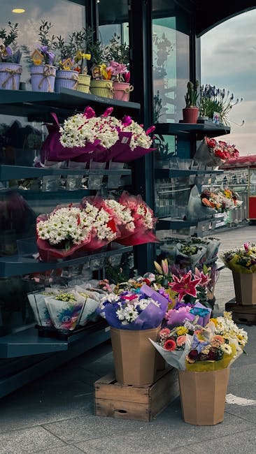 A display of fresh floral arrangements and bouquets arranged on black metal shelves outside a florist shop, featuring a variety of flowers in vibrant colors such as white, pink, purple, and yellow. Th