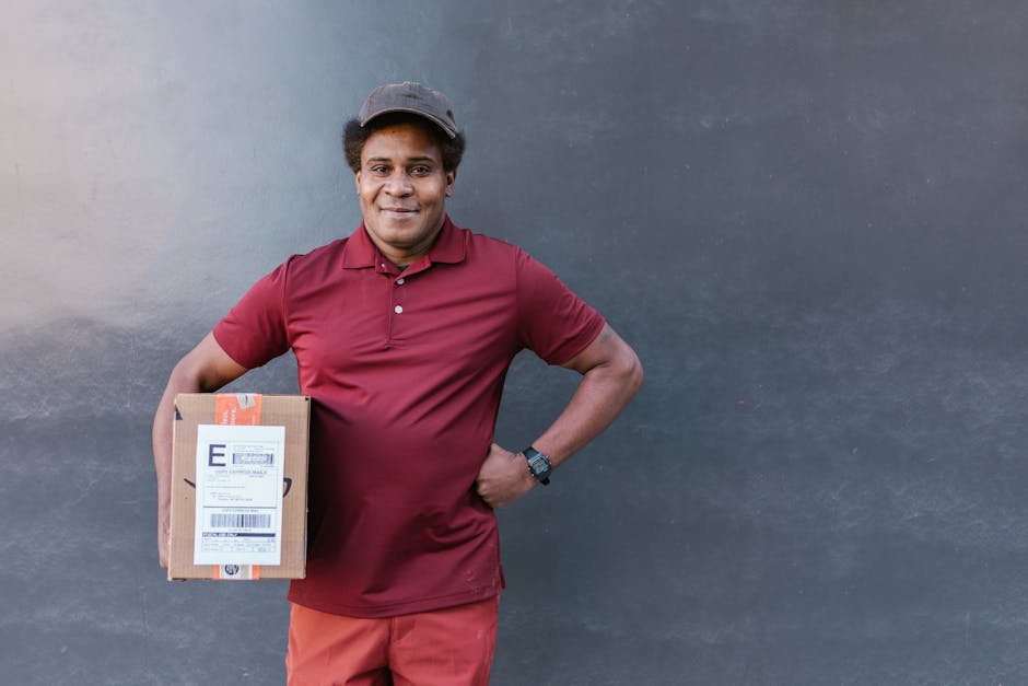 A smiling delivery person with dark skin, wearing a maroon polo shirt, matching maroon pants, and a dark cap, holding a small cardboard parcel with a shipping label attached. The person stands against