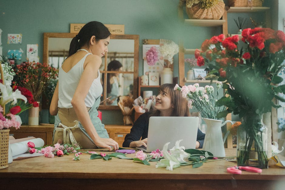 A young female florist and a female customer engage in a discussion inside a flower shop. The florist, wearing a light-colored sleeveless top and an apron, stands next to a large wooden table covered