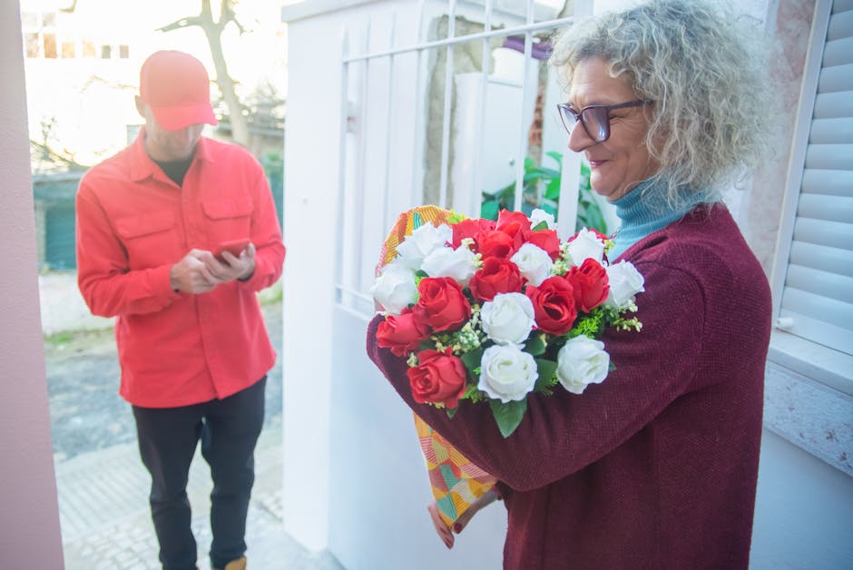 A woman with curly gray hair, wearing glasses, a blue turtleneck sweater, and a burgundy cardigan, is holding a vibrant bouquet of fresh red and white roses wrapped in colorful paper, ready for flower