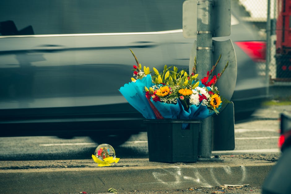 A vibrant bouquet of fresh flowers arranged with sunflowers, red and yellow blooms, wrapped in blue paper, and placed in a black container on the pavement at a street corner. The arrangement includes